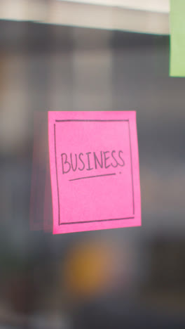 Vertical-Video-Close-Up-Of-Woman-Putting-Sticky-Note-With-Business-Written-On-It-Onto-Transparent-Screen-In-Office